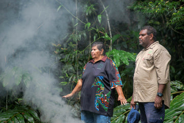 Two aboriginal people walking through a forest with smoke in the background