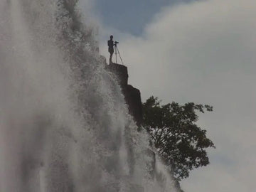Desre with a camera on a tripod standing on a rocky outcrop amidst a waterfall.