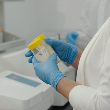 A person in a white lab coat wearing blue gloves holds a small clear specimen container containing skincare samples in a clinical laboratory setting.