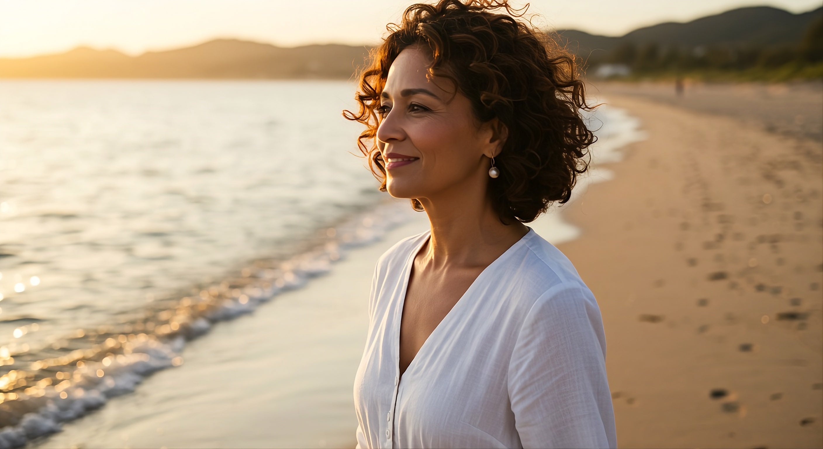 A smiling 60 year old woman in a white dress standing on a beach looking out to sea with mountains in the background.