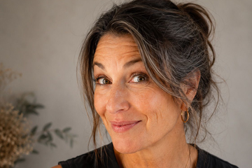 A close-up portrait of a woman with dark brown hair styled in a messy bun, wearing a black top and gold hoop earrings smiling mischievously at the camera.