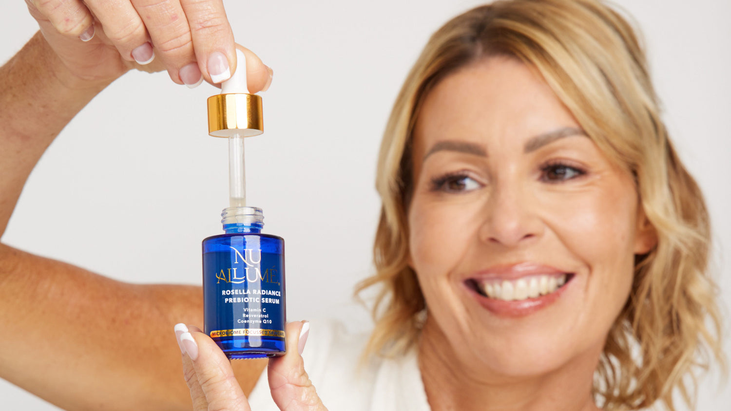 Middle aged woman smiling as she's holding a bottle of Nu Allumé Rosella Radiance Prebiotic Serum with a dropper.