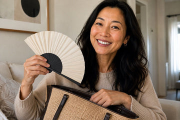 A smiling woman with long dark hair holds a black and cream folding hand fan next to a woven beige tote bag with black straps in a brightly lit living room.