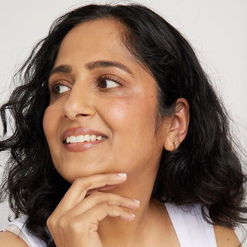A close-up portrait of a woman with dark wavy hair, wearing a white tank top and a small gold hoop earring, looking thoughtfully to the side against a light gray background.