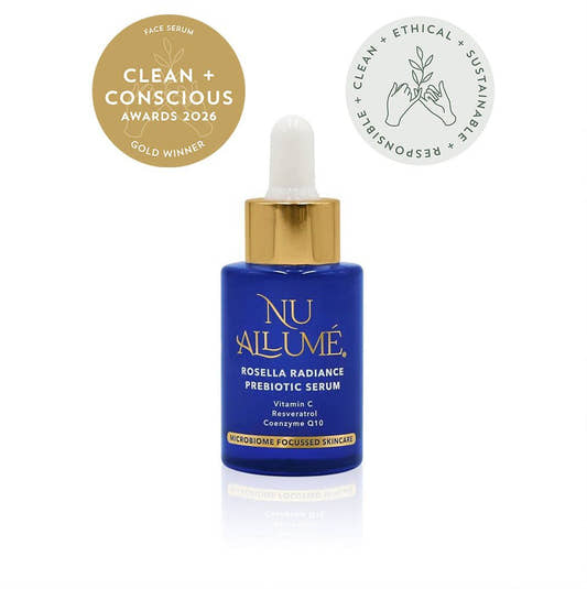 A blue dropper bottle of NU A LUMÉ Roscella Radiance Prebiotic Serum with a gold cap, shown on a white background with two award badges at the top: a gold 'Clean + Conscious Awards 2026 Gold Winner' seal and a white 'Clean + Ethical + Sustainable' seal.