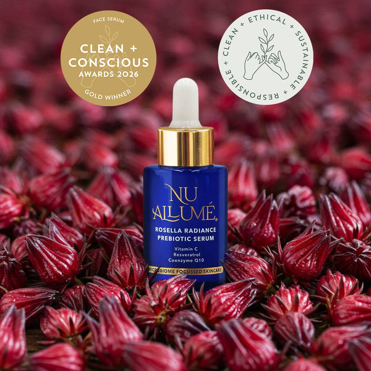 A blue and gold dropper bottle of NU AILUMÉ face serum stands centred among many red rosella flowers. Two award badges appear at the top: a gold circular badge reading 'Clean + Conscious Awards 2026 Gold Winner' and a white circular badge with sustainability text.