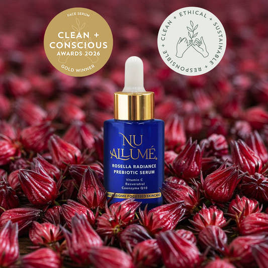 A blue and gold dropper bottle of NU AILUMÉ face serum stands centred among many red rosella flowers. Two award badges appear at the top: a gold circular badge reading 'Clean + Conscious Awards 2026 Gold Winner' and a white circular badge with sustainability text.