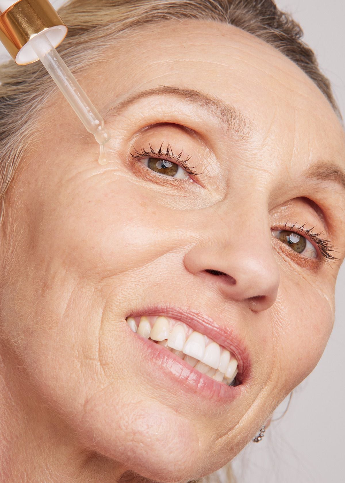 Close-up of a smiling woman applying a dropper of Nu Allume Rosella Radiance Prebiotic Serum to her face.