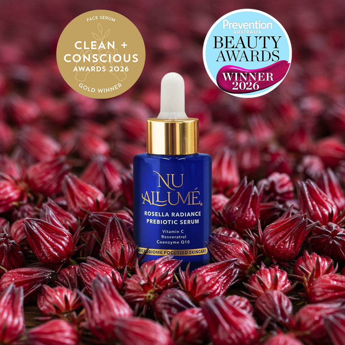 A blue and gold dropper bottle of NU AILUMÉ Rosella Radiance Prebiotic Serum is centred over a bed of red hibiscus flowers. Two award seals are displayed in the top corners: a gold 'Clean + Conscious Awards 2026 Gold Winner' seal on the left and a blue and pink 'Prevention Beauty Awards 2026 Winner' seal on the right.